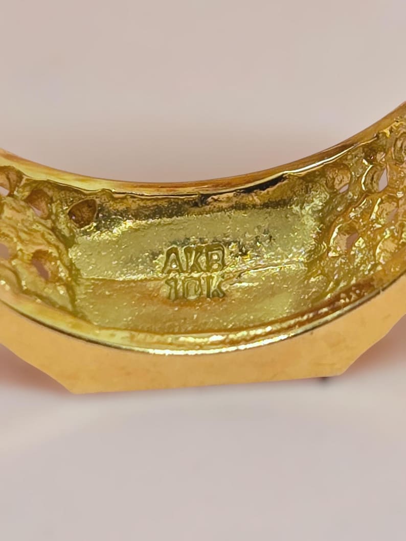 May include: A gold ring featuring a textured, porous surface on the sides and a smooth inner band. The interior of the ring is clearly stamped with the text AKB and 10K, indicating the metal purity and manufacturer mark.