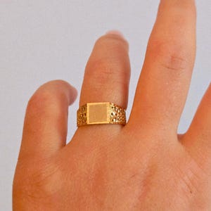 May include: A gold ring featuring a central square flat plate with a brushed finish, flanked by bands with a textured, perforated, or dimpled pattern. The metal has a warm, polished yellow tone throughout the entire band design.
