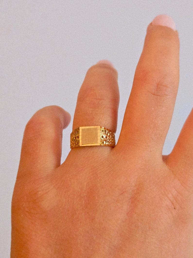 May include: A gold ring featuring a central square flat plate with a brushed finish, flanked by bands with a textured, perforated, or dimpled pattern. The metal has a warm, polished yellow tone throughout the entire band design.