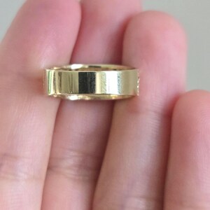 May include: A polished gold band ring with a flat, smooth exterior surface and a uniform width, showcasing a reflective metallic finish that catches the light across its entire circumference.