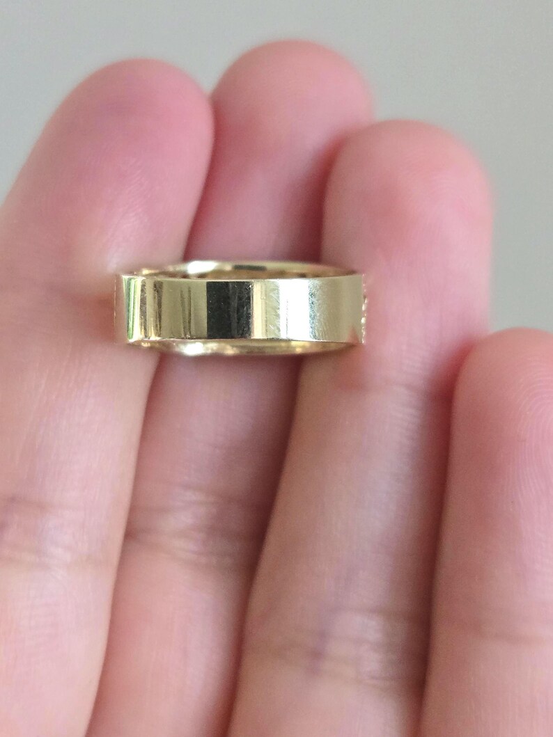 May include: A polished gold band ring with a flat, smooth exterior surface and a uniform width, showcasing a reflective metallic finish that catches the light across its entire circumference.