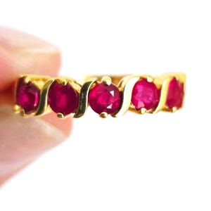 May include: A gold band ring featuring five round, vibrant red rubies set in a continuous row. The metal setting between each gemstone has a curved, S-shaped design that separates the stones, creating a fluid and connected appearance across the band.