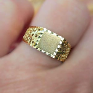 May include: A gold ring featuring a central rectangular flat plate with a brushed finish, flanked by textured, diamond-cut side panels that create a reflective, faceted appearance across the band's surface.
