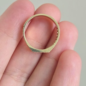 May include: A gold-toned ring featuring a flat, rectangular top surface and a smooth, circular band. The metal has a polished finish, and the design includes a series of small, evenly spaced indentations along the sides of the band.