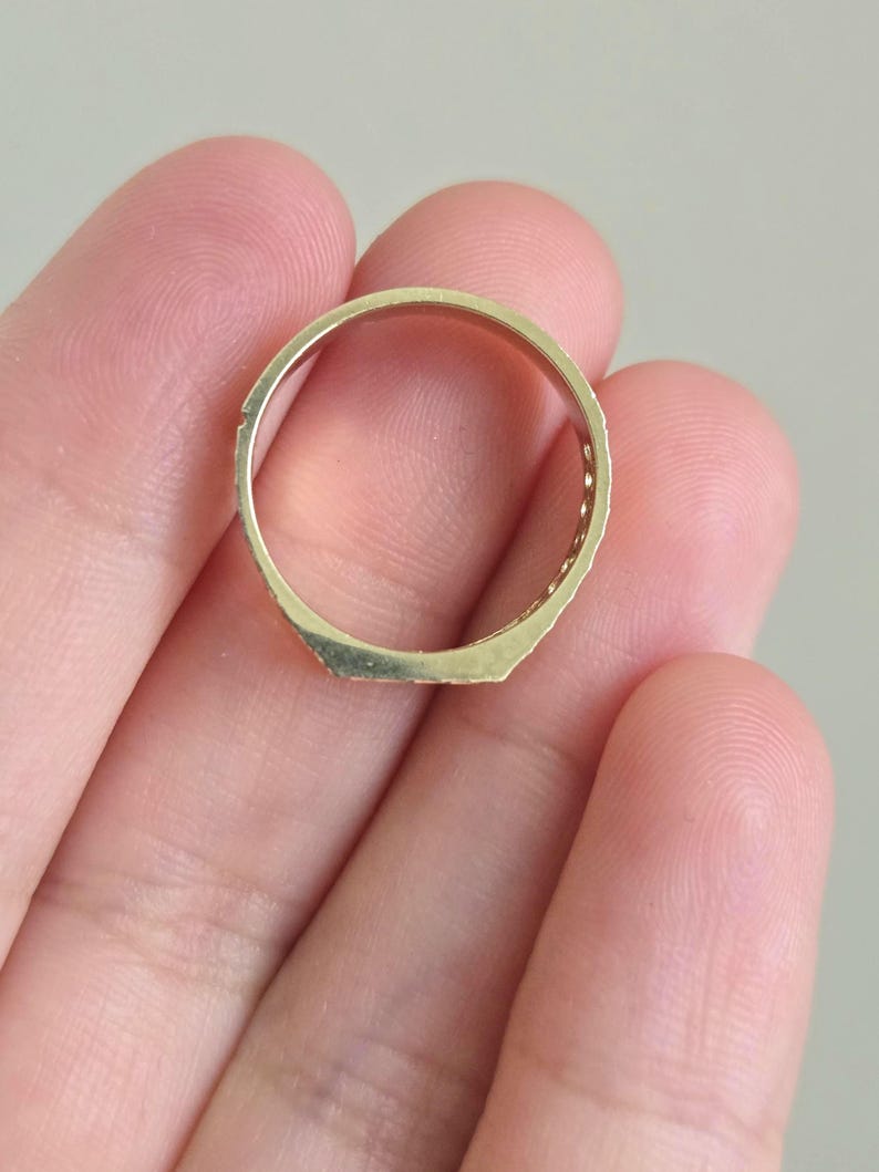 May include: A gold-toned ring featuring a flat, rectangular top surface and a smooth, circular band. The metal has a polished finish, and the design includes a series of small, evenly spaced indentations along the sides of the band.