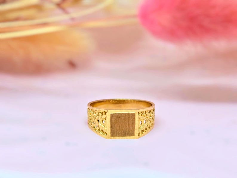 May include: A gold ring featuring a flat, rectangular center plate with a brushed finish. The band is constructed with an intricate, openwork filigree pattern that extends from the central plate around the sides of the ring.