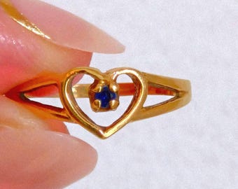 May include: A gold-colored ring featuring a heart-shaped design with a small, square, dark blue gemstone at its center. The ring has a split band design. The ring is held by a person.