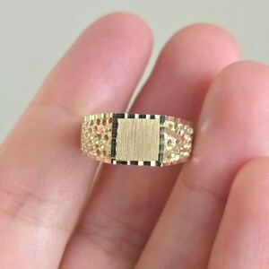 May include: A gold ring featuring a central square panel with a brushed finish, flanked by textured, openwork sides with a diamond-cut pattern. The band has a polished gold border and a detailed, intricate design across the entire surface.