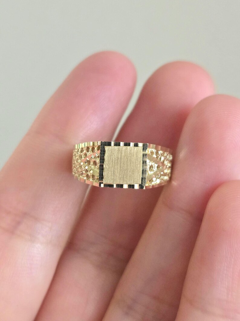 May include: A gold ring featuring a central square panel with a brushed finish, flanked by textured, openwork sides with a diamond-cut pattern. The band has a polished gold border and a detailed, intricate design across the entire surface.