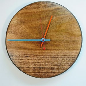 May include: A round wooden clock with a dark brown stain. The clock has a blue and orange hand. The clock is on a white wall.