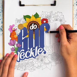 May include: A coloring page with a floral design and the words "do it recklessly" in a stylized font. The page is on a clipboard and a person is coloring the page with a black pencil.