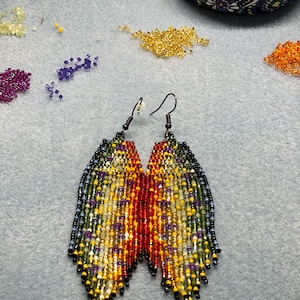 Puede incluir: Un par de pendientes de cuentas hechos a mano con un diseño de flecos. Los pendientes presentan un degradado de colores, incluyendo rojo, amarillo, verde y azul. Los pendientes están sobre una superficie azul claro, con cuentas dispersas en varios colores.