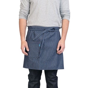 Puede incluir: Un delantal a rayas azules y blancas con un solo bolsillo. El delantal está atado a la cintura con una cinta azul. La persona que lleva el delantal lleva una camisa gris de manga larga y pantalones vaqueros azules.