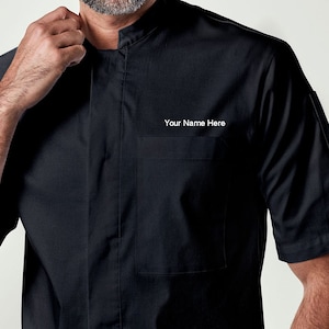 May include: Black short-sleeved chef jacket with a mandarin collar and a pocket on the left chest. The text "Your Name Here" is embroidered on the pocket.