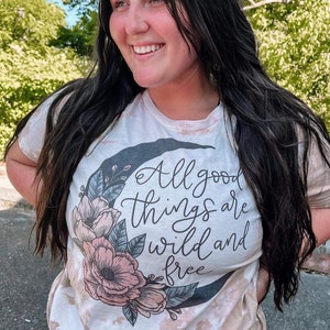 Könnte beinhalten: Ein weißes Batik-T-Shirt mit einem schwarzen Halbmond-Motiv. Der Mond ist von rosa und grünen Blumenillustrationen umgeben. Der Text "All good things are wild and free" ist in schwarzer Kursivschrift geschrieben.