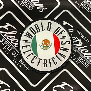 May include: A white, round patch with black text that reads "World of Electricians" and a lightning bolt symbol. The patch features the Mexican flag in the center.