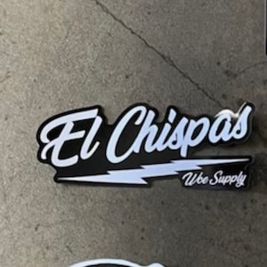 May include: Black and white sticker with the text "El Chispas" in a white cursive font with a white lightning bolt underneath. The text "Woe Supply" is in smaller white text below the lightning bolt.
