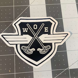 May include: Black and white sticker with a shield shape and a pair of crossed lacrosse sticks. The shield has wings and the text "WOE" in the center.