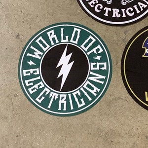 Könnte beinhalten: Ein runder Aufkleber in Grün und Weiß mit dem Text "World of Electricians" in einem kreisförmigen Design. Ein weißer Blitz befindet sich in der Mitte des Aufklebers.