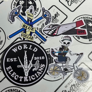 May include: A collection of stickers featuring electrician-themed designs. Includes a sticker with crossed blue pliers and the words "Blue Collar", a stylized shark-shaped tool, and a skeleton riding a bicycle. Other stickers have the text "World Electricians".