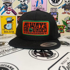 以下が含まれることがあります： 「Always Grounded」と「World of Electricians」と書かれたパッチが付いた黒と緑の野球帽。