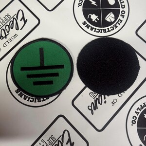 May include: Two circular patches, one featuring a green background with a black electrical ground symbol and a black border, and the other a solid black textured patch. Both items are round and display distinct contrasting colors and textures.