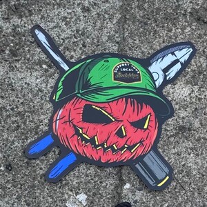 May include: A sticker featuring a red jack-o'-lantern wearing a green baseball cap with the text "SUPPORT YOUR LOCAL Electricians" on a patch. Behind the pumpkin are crossed blue and grey tools, including a screwdriver and pliers.