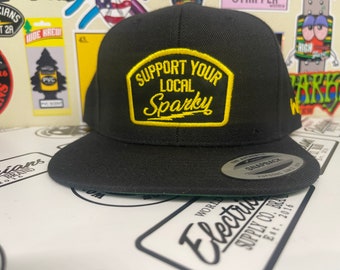 May include: Black baseball cap with a yellow patch that says "Support Your Local Sparky". The cap has a black brim and a black strap with a silver Snapback logo.