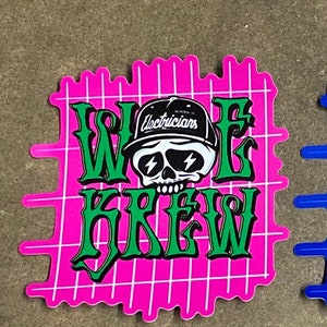 May include: A pink and green sticker with a skull wearing a black hat that says "Electricians". The sticker says "WOLF KREW" in green letters on a pink grid background.