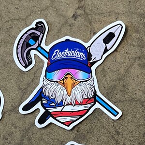 Könnte beinhalten: Ein Cartoon-Glatzkopfadler trägt eine blaue Baseballkappe mit dem Text "World of Electricians" und eine rosafarbene Skibrille. Der Adler trägt ein rot-weiß-blaues Bandana. Der Adler hält einen Schraubenschlüssel und einen Hammer.