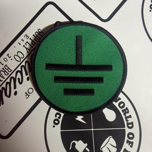 May include: A circular embroidered patch featuring a vibrant green background with a black electrical ground symbol centered on the fabric. The patch has a thick black stitched border that defines its round shape and provides a clean finish.