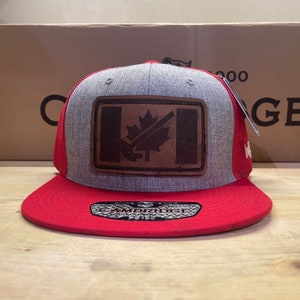 Könnte beinhalten: Eine rote und graue Baseballkappe mit einem braunen Lederpatch, auf dem eine kanadische Flagge mit einem Ahornblatt abgebildet ist. Die Kappe hat ein Netzrücken und der Text "CAMBRIDGE" ist auf der Vorderseite sichtbar.