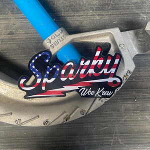 May include: A black and red sticker with the word "Sparky" in white lettering and the words "Woe Kreu" below. The sticker has an American flag design and is attached to a silver metal pipe bender.