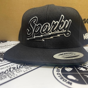 May include: Black and white baseball cap with the word "Sparky" embroidered on the front in a white outline with a black fill. The cap has a black snap closure and a white "World of Electric" logo on the side.