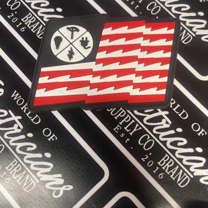 May include: A red, white, and black patch with a stylized American flag design. The flag has lightning bolts instead of stars. A circle in the upper left corner contains symbols related to electricity: a light bulb, a plug, a power cord, and a lightning bolt.