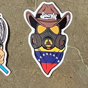 May include: A sticker featuring a cartoon character wearing a brown cowboy hat with the word "Sparky" on it, a gas mask, and a bandana with the Venezuelan flag.