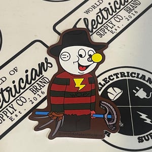 May include: A sticker featuring a cartoon character with a red and black striped shirt, a black hat, and a yellow light bulb on his head. The character is holding a blue and silver tool. The sticker has the text "woe" and "@worldofelectricians" on it.