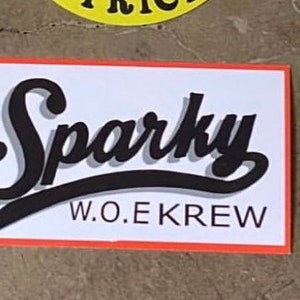 May include: A white sticker with an orange border features a black stylized logo that reads "Sparky" with the text "W.O.E KREW" below it.