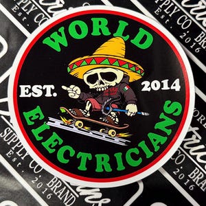 May include: A round sticker with a black background and a red border. The sticker features a cartoon skeleton wearing a sombrero and riding a skateboard. The text "WORLD ELECTRICIANS EST. 2014" is written in white letters around the skeleton.