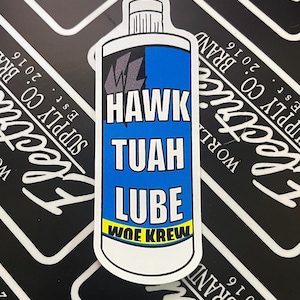 May include: A sticker featuring a blue and white bottle design with the words "HAWK TUAH LUBE" in bold white letters. The sticker also includes the text "WOE KREW" and a logo. The background is black and white.