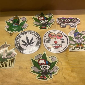 May include: A collection of colorful stickers with various cartoon characters and cannabis leaf designs. Some stickers feature text like "WORLD OF ELECTRICIANS" and "HUMBOLDT". The stickers are on a light brown wooden surface.