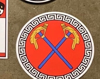May include: A red circle with a black and white Greek key border. Inside the circle are two crossed golden axes with blue handles.
