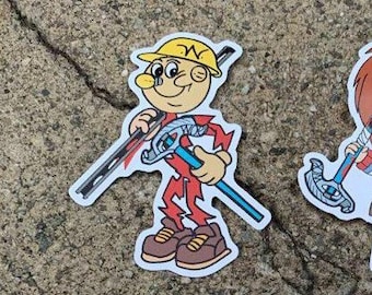 May include: A cartoon sticker of a worker wearing a yellow hard hat and red overalls holding a wrench and a pipe. The sticker is on a gray concrete surface.