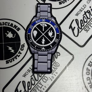 Peut inclure: Un autocollant en forme de montre avec une lunette bleue et noire. Le cadran de la montre présente des symboles électriques et le texte "WORLD ELECTRIC SUPPLY CO."