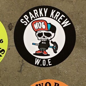May include: A black and white circular sticker with the text "Sparky Krewe" and "W.O.E."  A cartoon skull wearing a red, white, and blue baseball cap with the text "W.O.E." is in the center of the sticker.