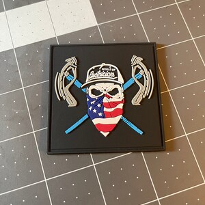 Könnte beinhalten: Ein schwarzer und blauer Patch mit einem Schädel, der eine Baseballkappe mit der Aufschrift "World of Electricians" trägt. Der Schädel trägt ein Bandana mit dem Design der amerikanischen Flagge. Der Schädel hält zwei gekreuzte Schraubenschlüssel.