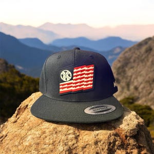 May include: A black baseball cap with a red, white, and blue American flag patch. The flag has a stylized lightning bolt design and a circular patch with a compass, axe, and campfire graphic. The cap has a black brim and a white "SNAPBACK" label.