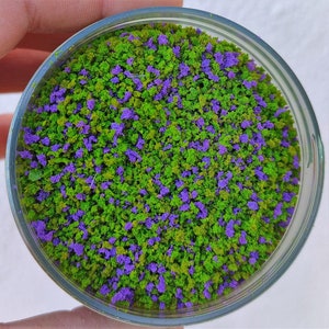 以下が含まれることがあります： 緑と紫のマイクロビーズが混ざったガラス瓶のクローズアップ。ビーズは、小さな緑の木々と紫の花が咲くミニチュアの森に似ています。