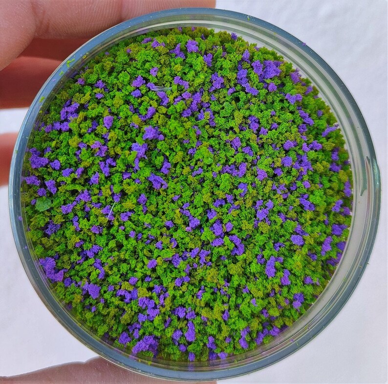 以下が含まれることがあります： 緑と紫のマイクロビーズが混ざったガラス瓶のクローズアップ。ビーズは、小さな緑の木々と紫の花が咲くミニチュアの森に似ています。