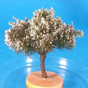 May include: A small, white-flowered tree model with a brown trunk and a base made of brown and yellow material. The tree is set against a blue background.